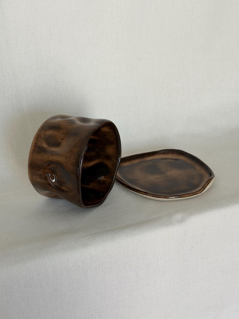 Mottled brown cup and saucer, a handbuilt ceramic cup pressed into dimples under a mottled brown glaze, 10 oz